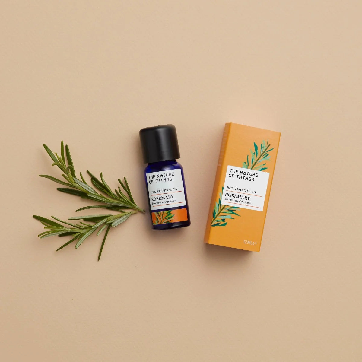 Kleine blaue 12-ml-Flasche des Essential Oil Organic Rosemary von The Nature of Things und dazugehörige orangefarbene Umverpackung mit Rosmarin-Illustrationen vor einem beigen Hintergrund.