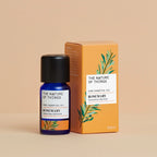 Kleine blaue 12-ml-Flasche des Essential Oil Organic Rosemary von The Nature of Things und dazugehörige orangefarbene Umverpackung mit Rosmarin-Illustrationen vor einem beigen Hintergrund.