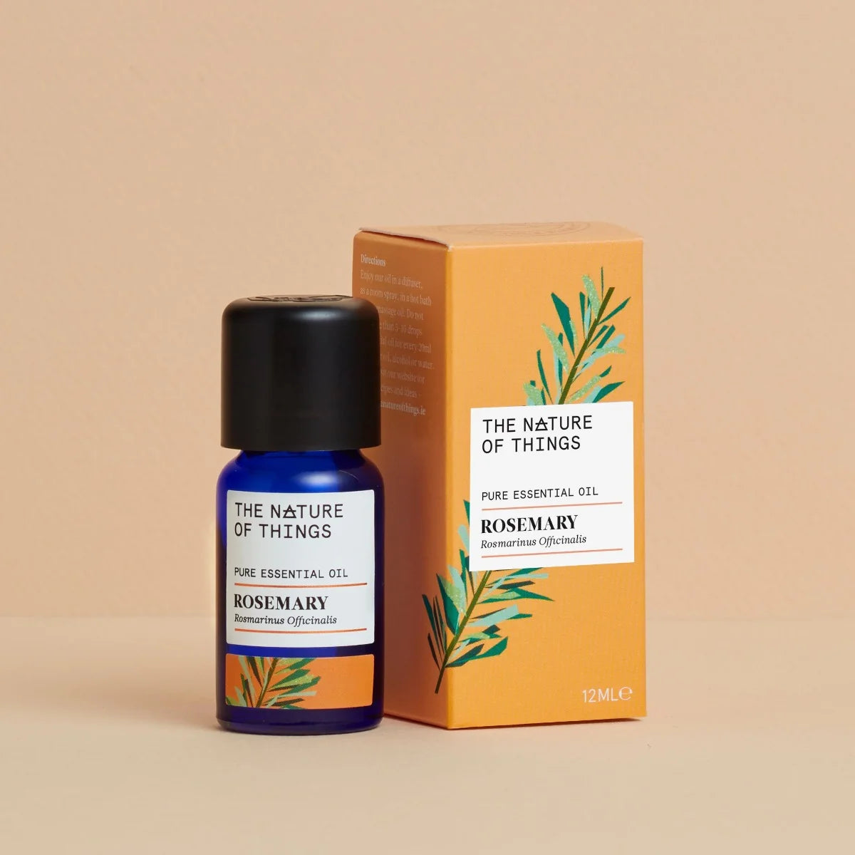 Kleine blaue 12-ml-Flasche des Essential Oil Organic Rosemary von The Nature of Things und dazugehörige orangefarbene Umverpackung mit Rosmarin-Illustrationen vor einem beigen Hintergrund.