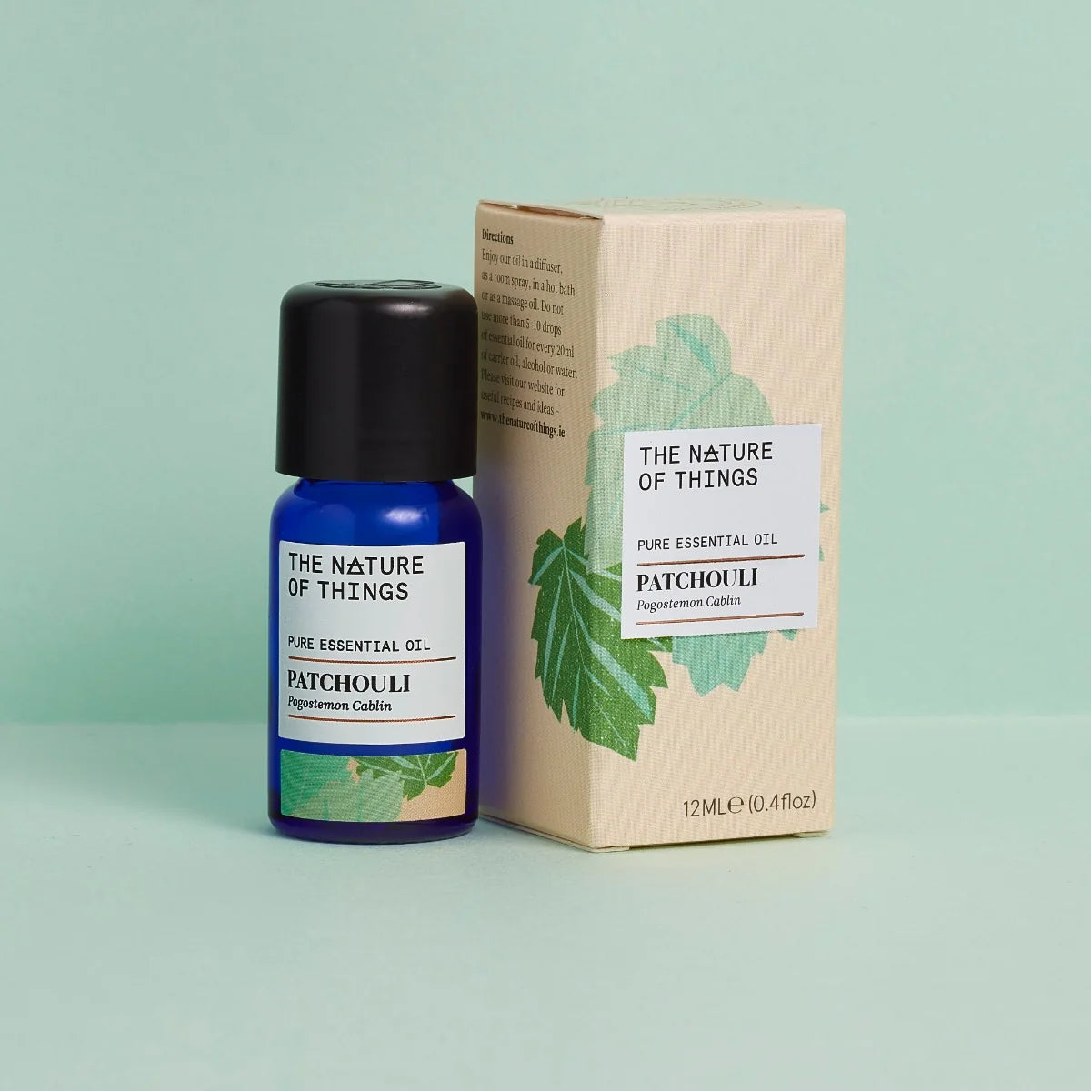 Kleine blaue 12-ml-Flasche des Essential Oil Patchouli von The Nature of Things und dazugehörige beige Umverpackung mit grünen Blatt-Illustrationen vor einem hellgrünen Hintergrund.