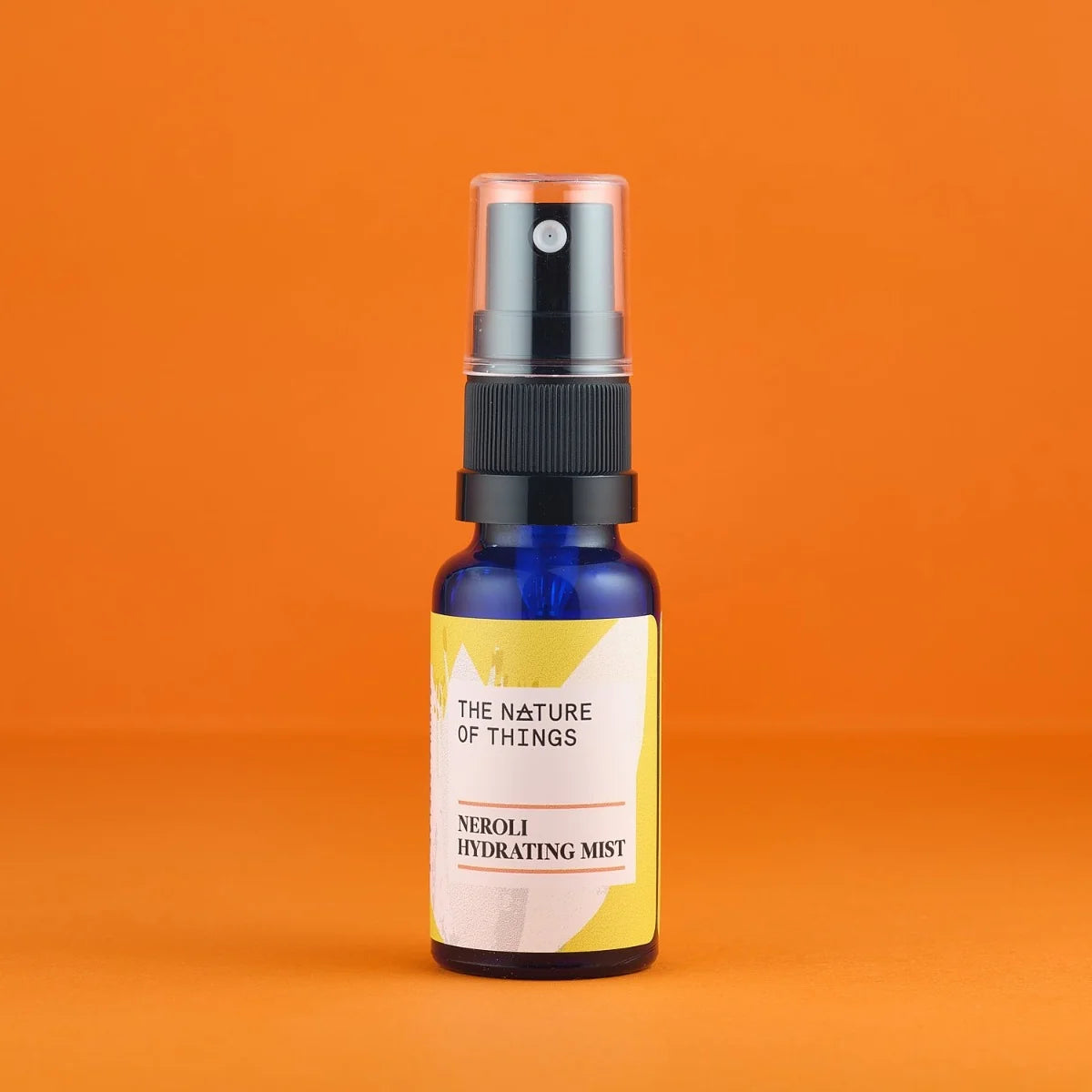 Kleine blaue 20ml-Sprühflasche des Neroli Mists von The Nature of Things mit gelb-weißem Etikett und durchsichtigem Sprühkopf auf orangefarbenem Hintergrund.