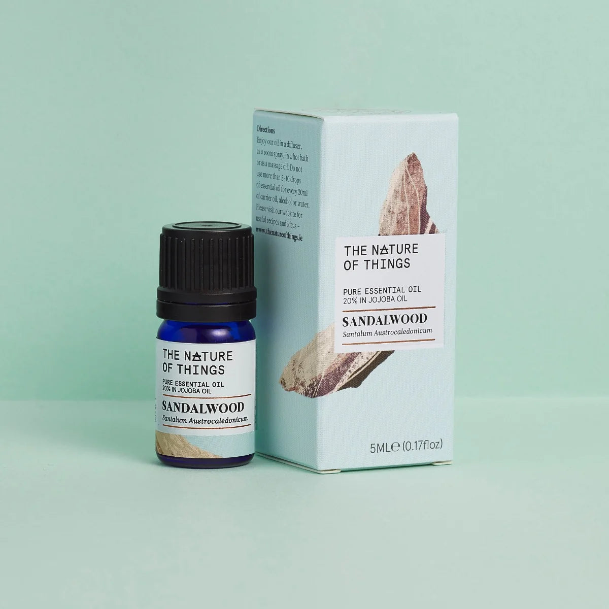 Kleine blaue 5-ml-Flasche des Essential Oil Sandalwood von The Nature of Things und dazugehörige hellblaue Umverpackung mit Holzdarstellung vor einem grünen Hintergrund.
