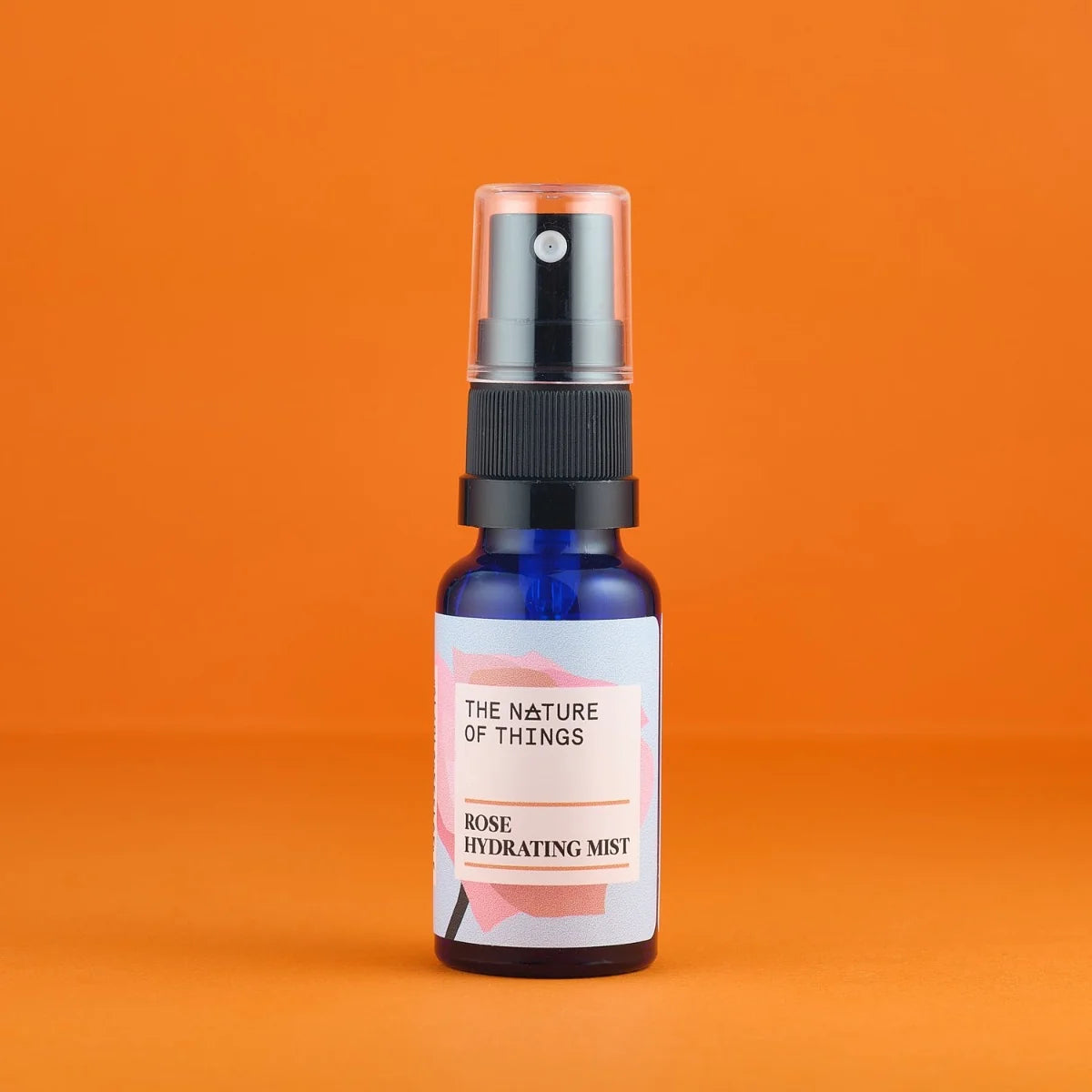 Kleine blaue Sprühflasche des Rose Mists von The Nature of Things mit rosa-weißem Etikett und durchsichtigem Sprühkopf auf orangefarbenem Hintergrund.