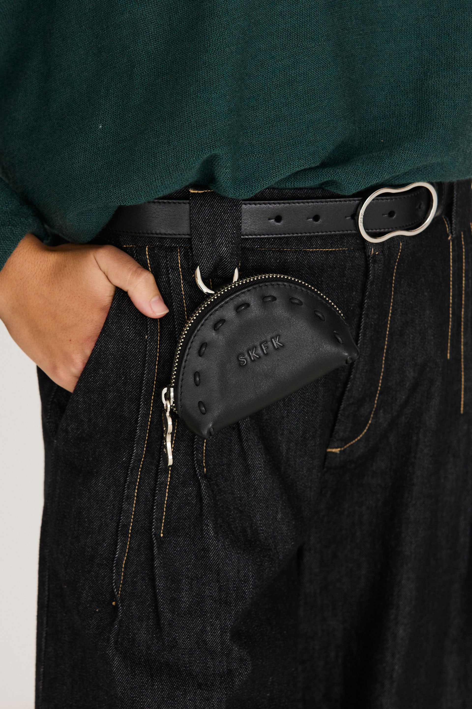 Kleine schwarze Geldbörse "Nea" mit geprägtem SKFK-Logo und Karabiner, befestigt an einer Gürtelschlaufe über einer dunklen Jeans.