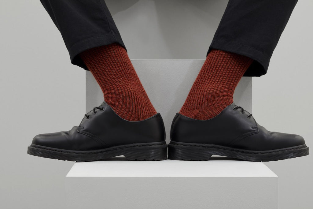 Ein Paar dicke, rustikal gestrickte Socken in Rostrot mit einem gerippten Bündchen. Die Bündchen haben ein Muster aus rostroten und braunen Streifen. Die Socken sind seitlich liegend vor einem weißen Hintergrund dargestellt.