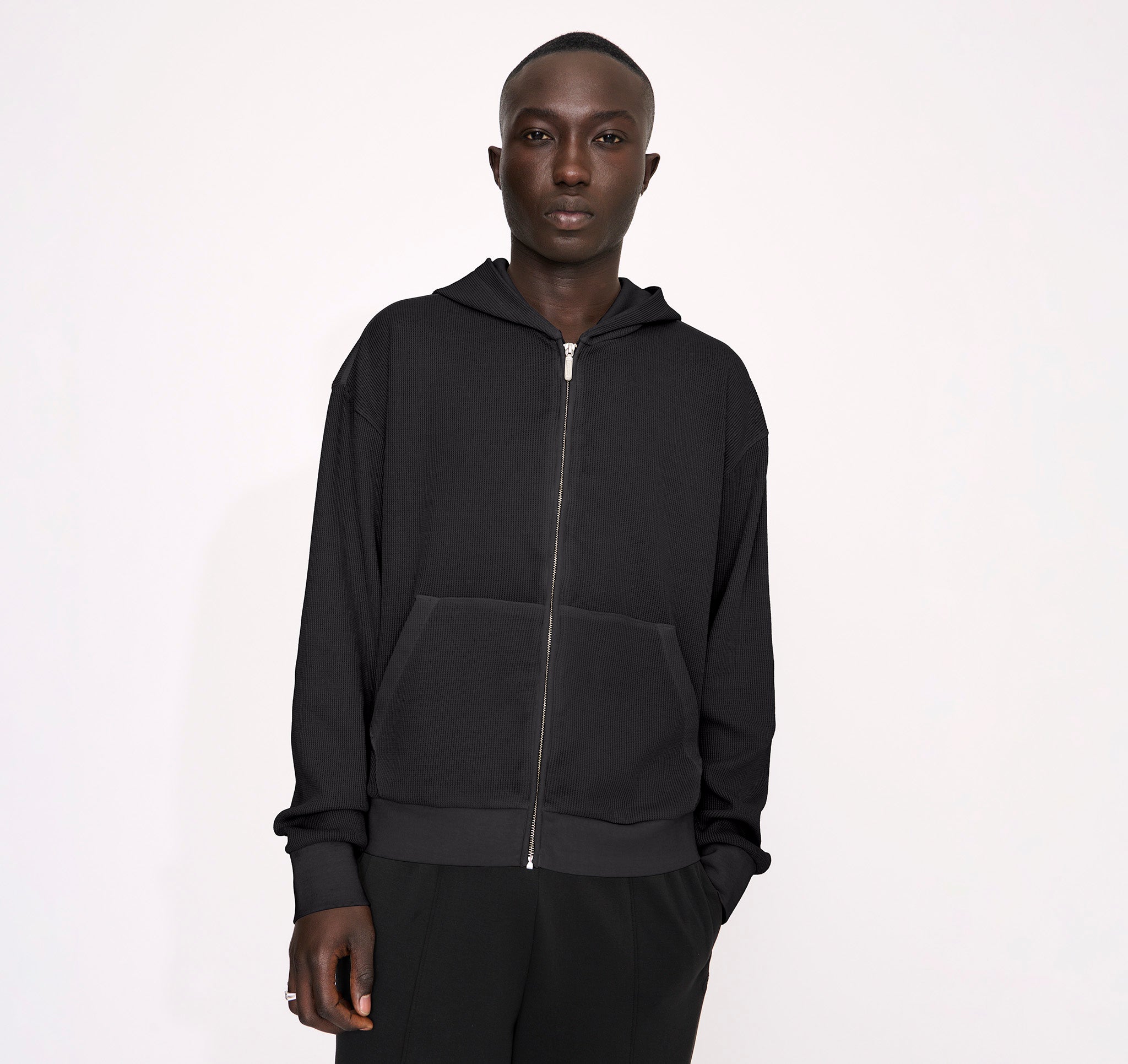 Männliches Model trägt einen dunkelgrauen Waffle Zip Hoodie mit strukturierter Waffeloptik und durchgehendem Reißverschluss vor hellem Hintergrund.