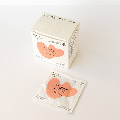 Minimalistische, quadratische weiße Teebox von rhoeco für den "Organic Cacao Husk Tea Original", verziert mit einer abstrakten pfirsichfarbenen Form und englischen sowie griechischen Produktinfos.