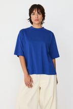 Model trägt das blaue Zuntz-11-Gots T-Shirt der Marke SKFK mit Stehkragen und kurzen Ärmeln zu hellen Hosen.