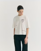 Model trägt das weiße Tomatera White Lena Tee von Thinking Mu mit detailreicher Stickerei eines Esels und Tomaten auf der Brust zu dunkler Hose.