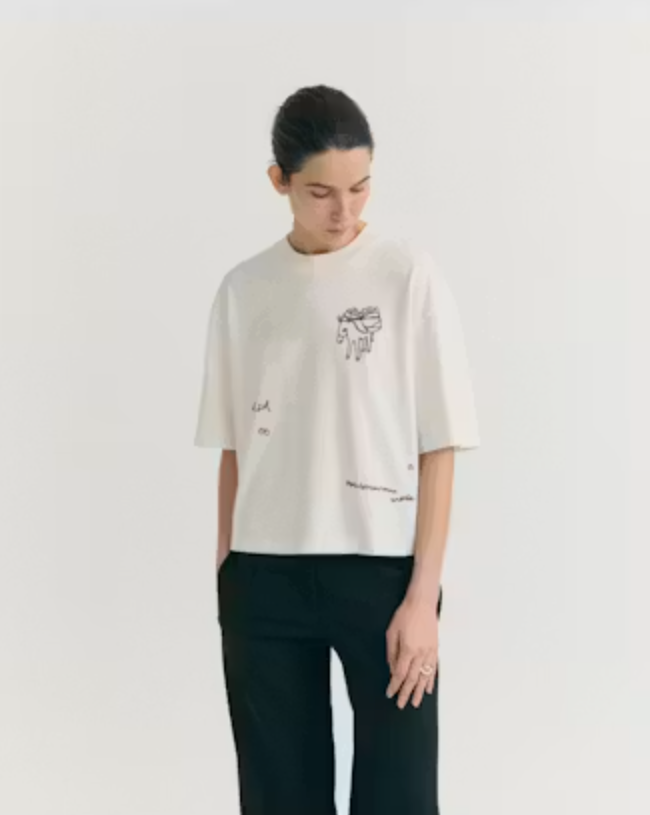 Model trägt das weiße Tomatera White Lena Tee von Thinking Mu mit detailreicher Stickerei eines Esels und Tomaten auf der Brust zu dunkler Hose.
