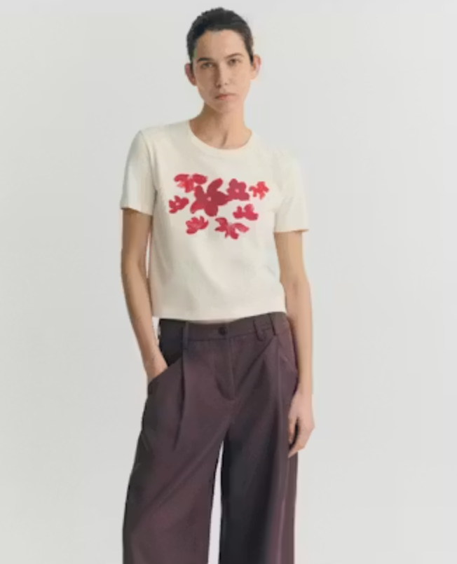 Model trägt ein cremeweißes Red Garden Pannacota Zowie Tee von Thinking Mu mit einem roten floralen Blumenmuster auf der Brust.