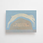 "No Road is too long" im DIN-A6-Format von Anna Cosma mit einem springenden weißen Hasen über eine Hügellandschaft vor blauschattiertem Hintergrund und dem Text „No road is too long in the company of a friend.“.