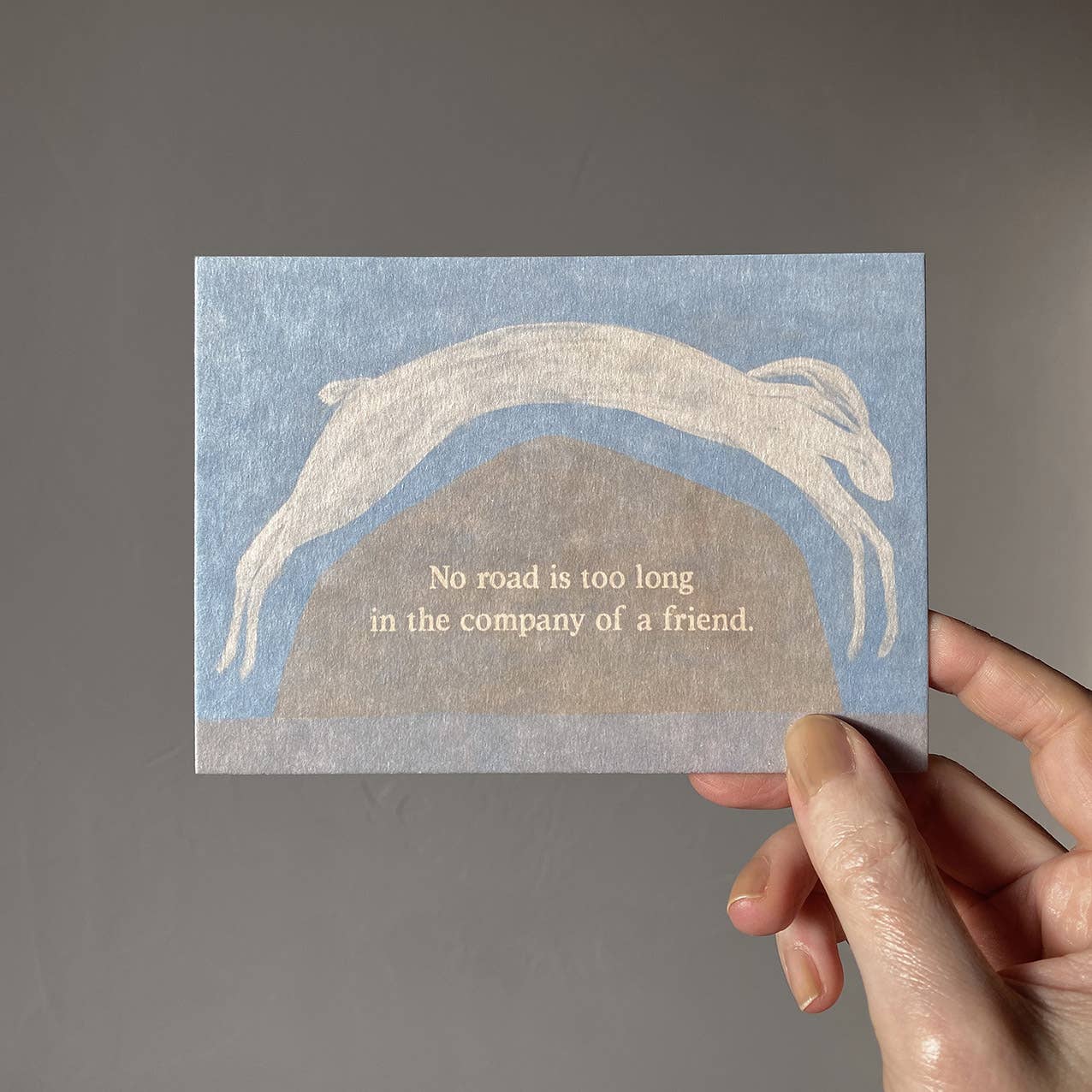 "No Road is too long" im DIN-A6-Format von Anna Cosma mit einem springenden weißen Hasen über eine Hügellandschaft vor blauschattiertem Hintergrund und dem Text „No road is too long in the company of a friend.“.