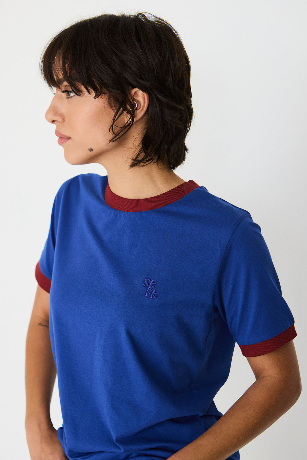 Person im Profil trägt ein blaues T-Shirt "Neskur" mit burgunderroten Kontrastbündchen und gesticktem SKFK-Logo vor hellem Hintergrund.