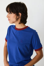 Person im Profil trägt ein blaues T-Shirt "Neskur" mit burgunderroten Kontrastbündchen und gesticktem SKFK-Logo vor hellem Hintergrund.