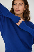 Person von vorne in einem blauen Strickpullover "Ibar" mit runden Halsausschnitt und Dolman-Ärmeln vor hellem Hintergrund.