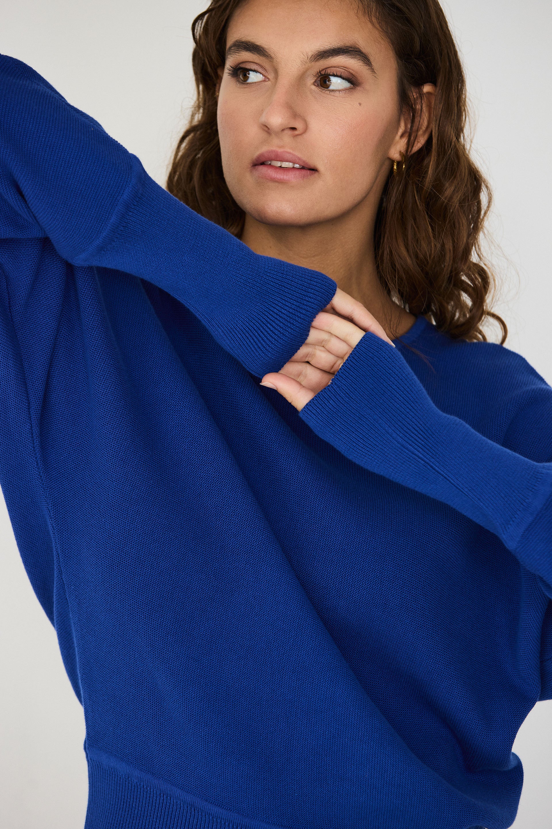 Person von vorne in einem blauen Strickpullover "Ibar" mit runden Halsausschnitt und Dolman-Ärmeln vor hellem Hintergrund.