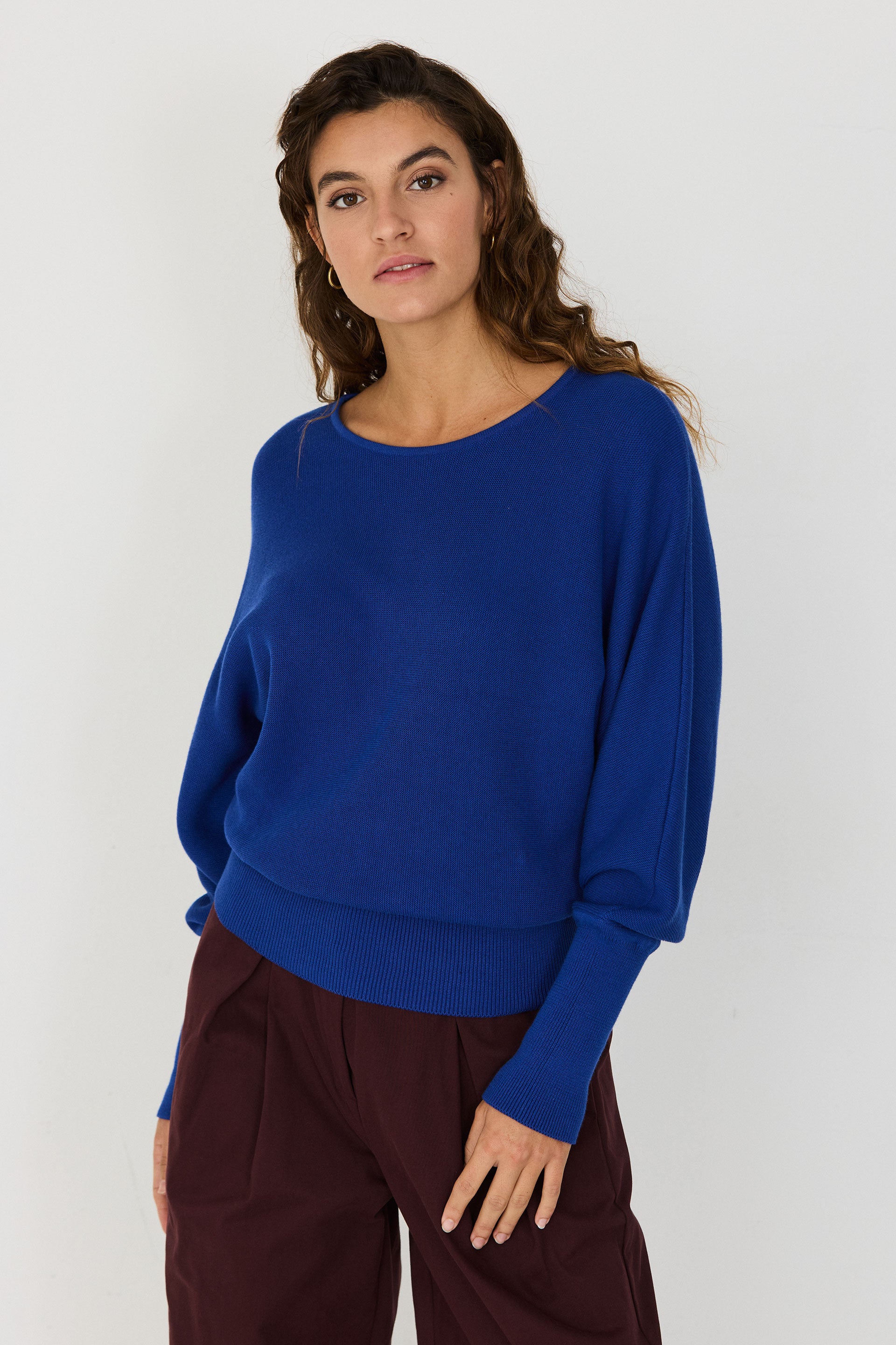 Person von vorne in einem blauen Strickpullover "Ibar" mit runden Halsausschnitt und Dolman-Ärmeln vor hellem Hintergrund.
