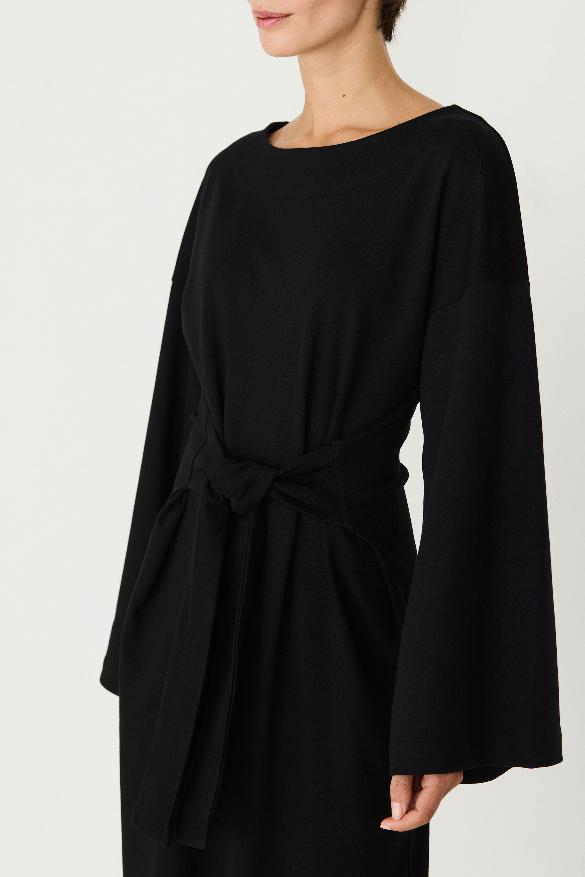 Person von vorne in einem schwarzen Langarmkleid "Domeka" mit Taillenbindung und weiten Kimonoärmeln vor hellem Hintergrund.