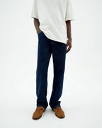 Person von vorne in einer dunklen Straight-Leg Jeans "David" mit hellem Shirt und braunen Schuhen vor hellem Hintergrund.