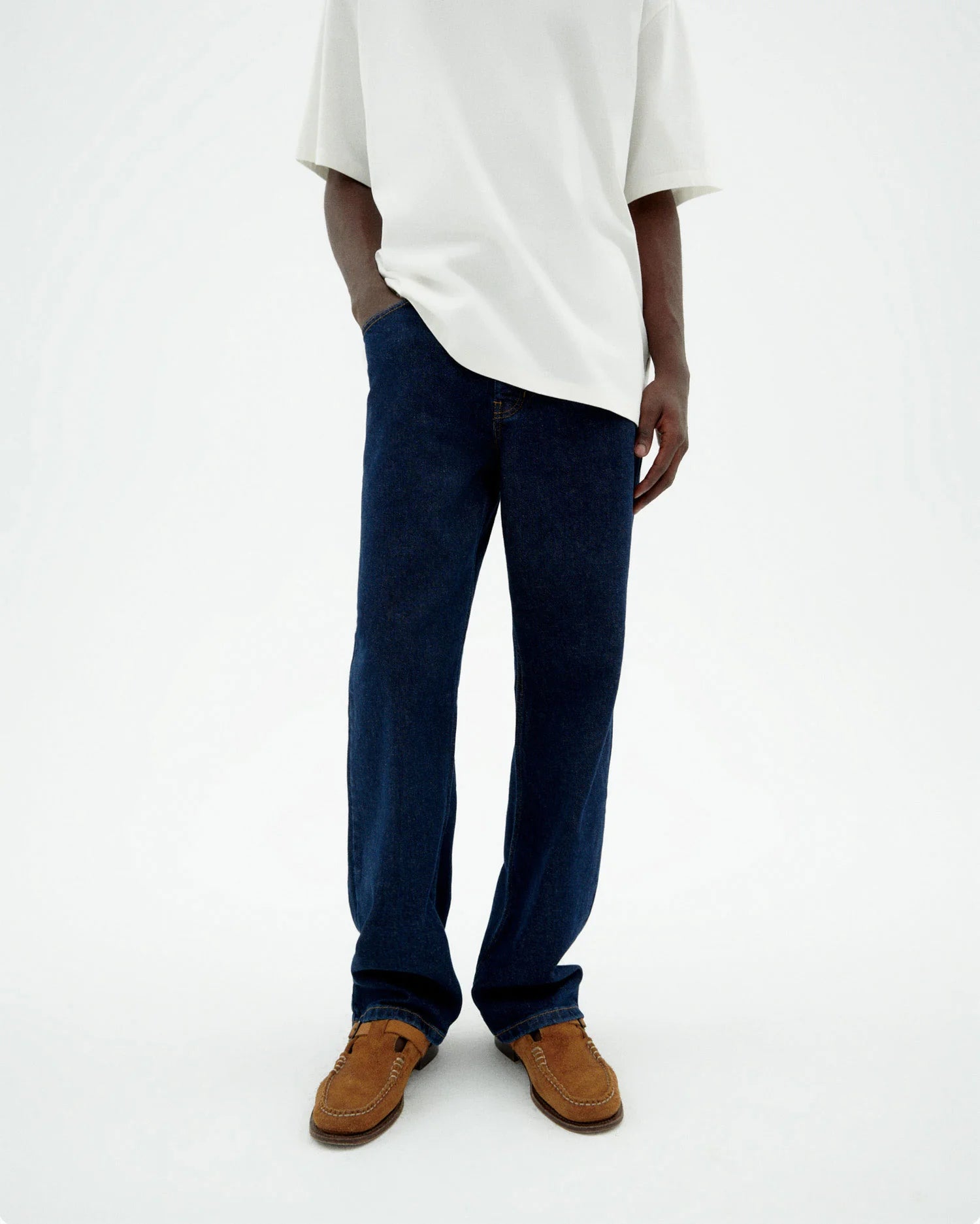 Person von vorne in einer dunklen Straight-Leg Jeans "David" mit hellem Shirt und braunen Schuhen vor hellem Hintergrund.