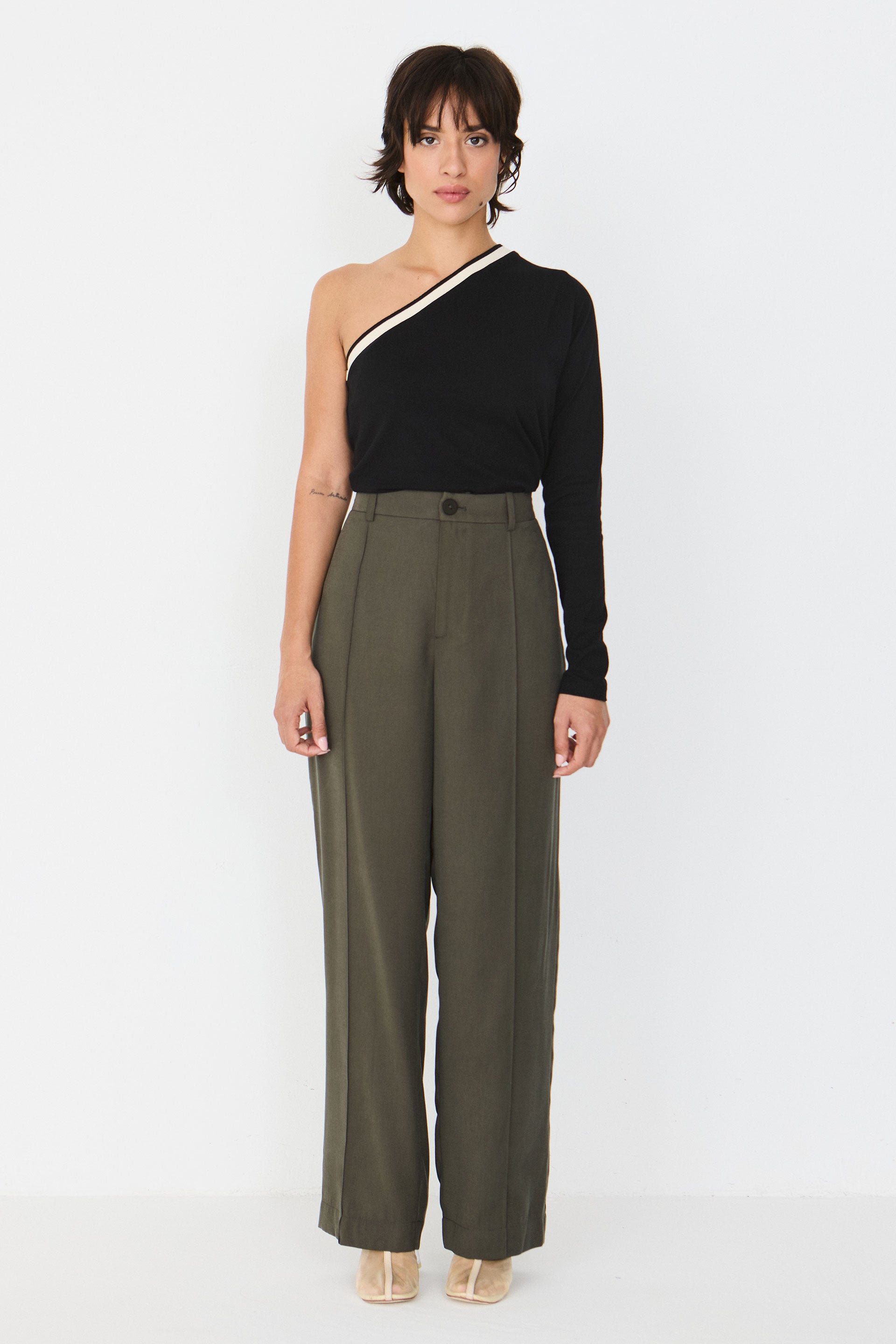 Person von vorne in einer geraden taupefarbenen Hose "Afrika" und einem schwarzen One-Shoulder-Oberteil vor hellem Hintergrund.