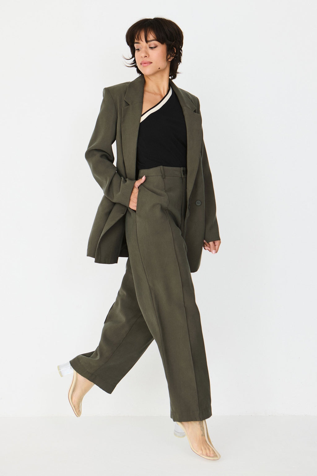 Person von vorne in einer geraden taupefarbenen Hose "Afrika" und einem schwarzen One-Shoulder-Oberteil vor hellem Hintergrund.