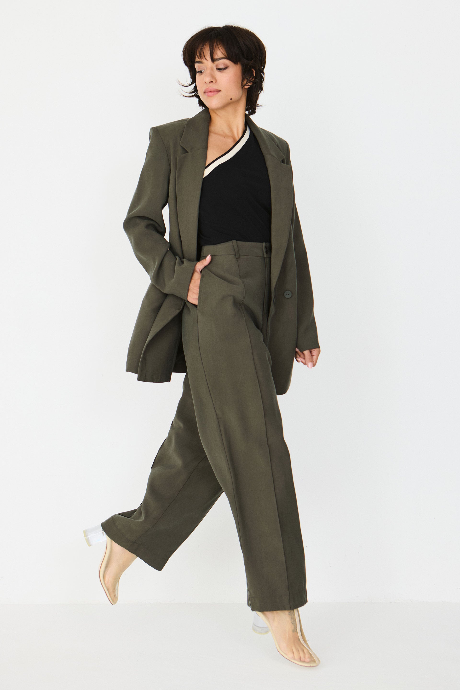 Person von vorne in einer geraden taupefarbenen Hose "Afrika" und einem schwarzen One-Shoulder-Oberteil vor hellem Hintergrund.