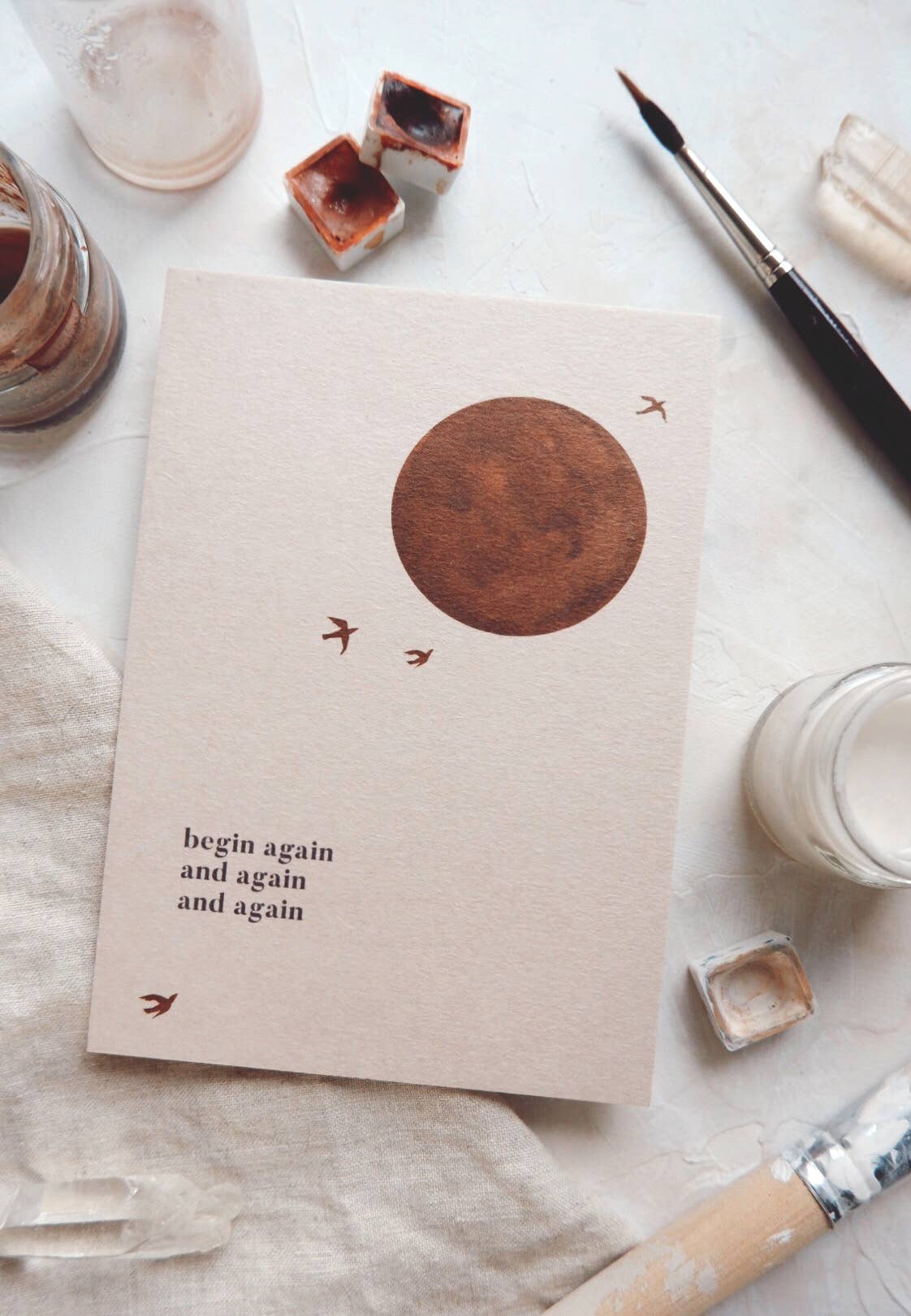 Postkarte "Begin Again" im DIN A6 Format von Anna Cosma mit einem runden braunen Motiv, kleineren Vögeln und dem Text "begin again and again and again" auf beigem Hintergrund.