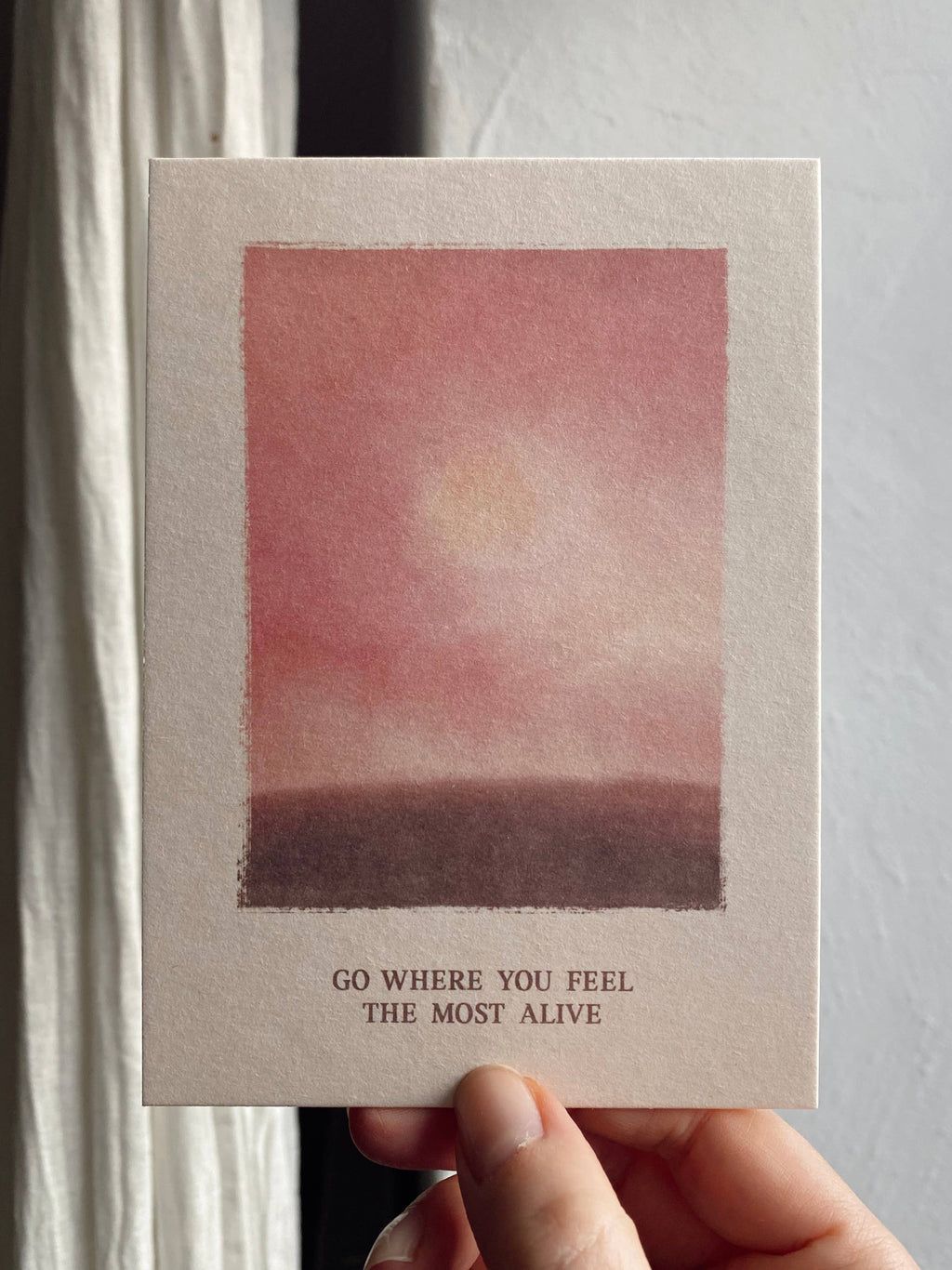 Postkarte „Go where you feel the Most Alive“ im DIN-A6-Format mit einer stimmungsvollen Illustration in Rosé-Tönen, einer sanften Sonne und dem Schriftzug vor hellem Hintergrund.