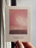 Postkarte „Go where you feel the Most Alive“ im DIN-A6-Format mit einer stimmungsvollen Illustration in Rosé-Tönen, einer sanften Sonne und dem Schriftzug vor hellem Hintergrund.