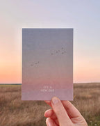 Postkarte „It's a New Day“ im DIN-A6-Format mit einem stimmungsvollen Farbverlauf, fliegenden Vögeln und dem Schriftzug vor einer natürlichen Landschaft.
