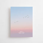 Postkarte „It's a New Day“ im DIN-A6-Format mit einem stimmungsvollen Farbverlauf, fliegenden Vögeln und dem Schriftzug vor einer natürlichen Landschaft.
