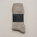 Zusammengelegte melierte Stricksocken aus Merinowolle und Seide im Naturton mit schwarzer Ferdinand-Banderole vor hellem Hintergrund.