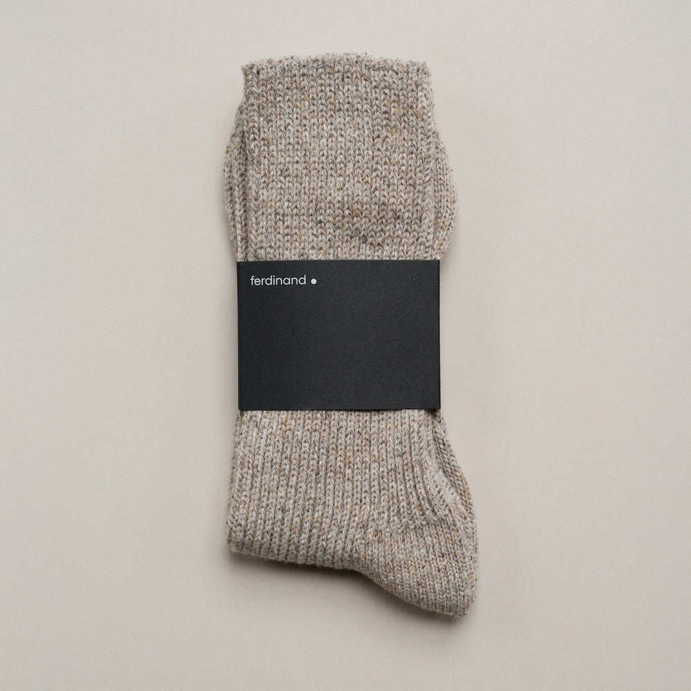 Zusammengelegte melierte Stricksocken aus Merinowolle und Seide im Naturton mit schwarzer Ferdinand-Banderole vor hellem Hintergrund.