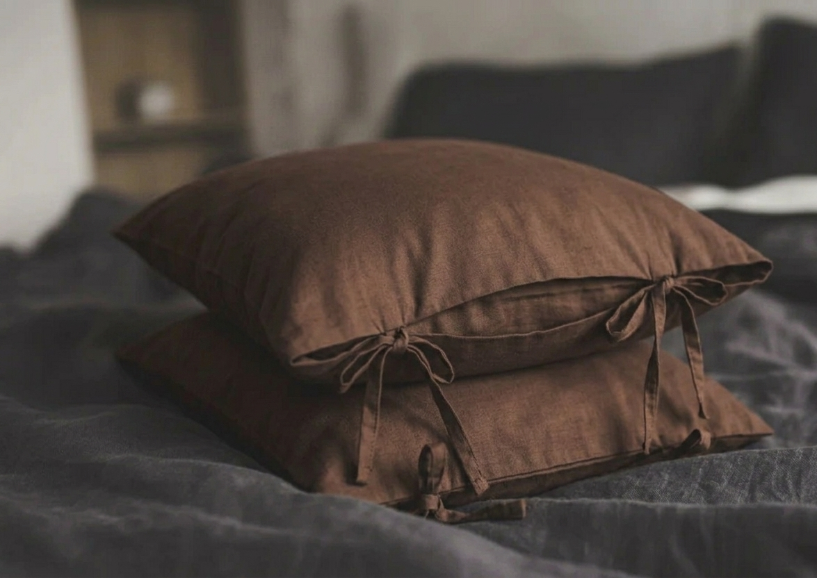 Zwei übereinander gestapelte, schokoladenbraune (Chocolate) Kissenbezüge aus edlem Leinenstoff mit dekorativen seitlichen Schleifenverschlüssen auf einem Bett.