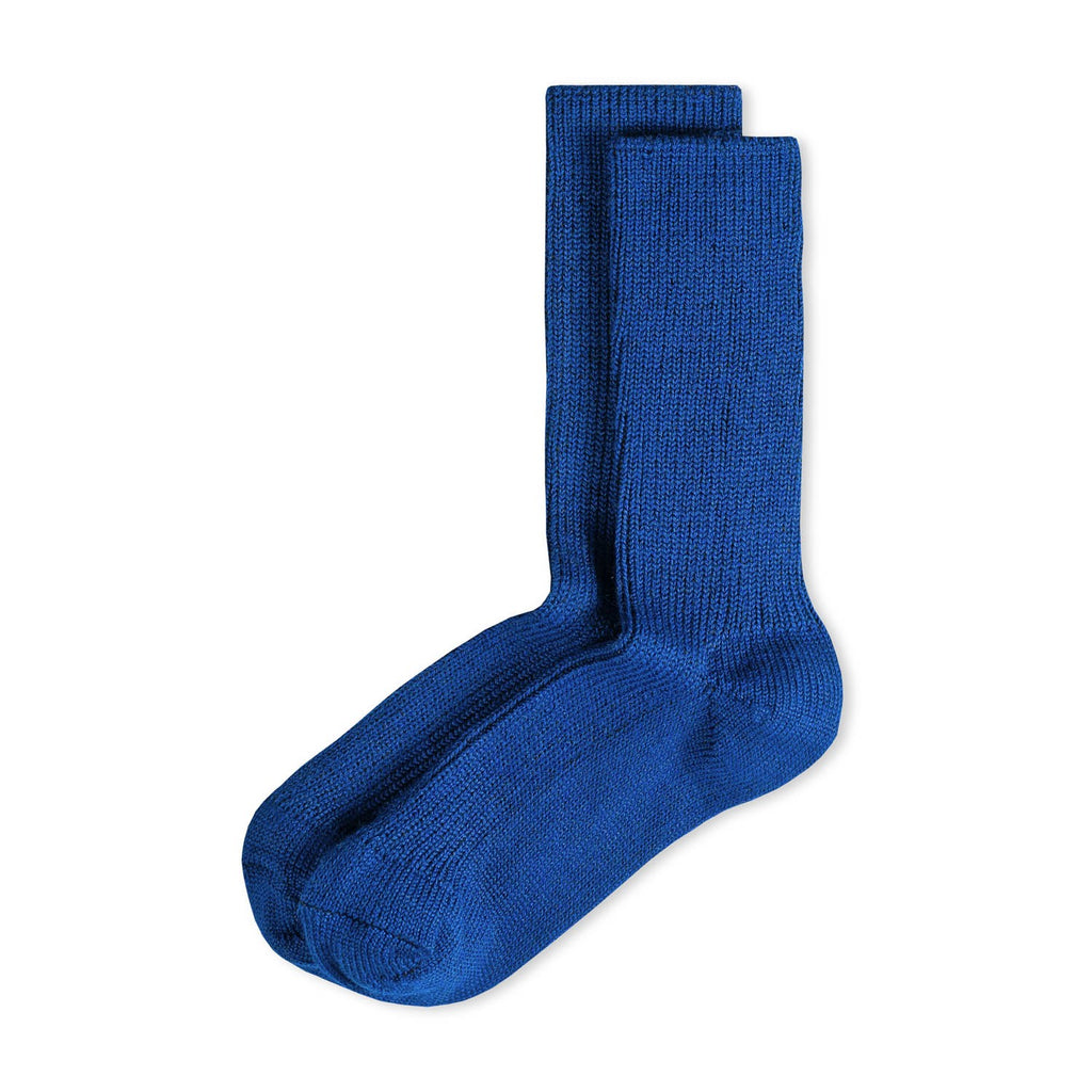 Zwei royalblaue Rippstricksocken mit hohem Schaft an den Füßen einer Person in schwarzer Hose, auf einer weißen Sitzfläche vor grauem Hintergrund.