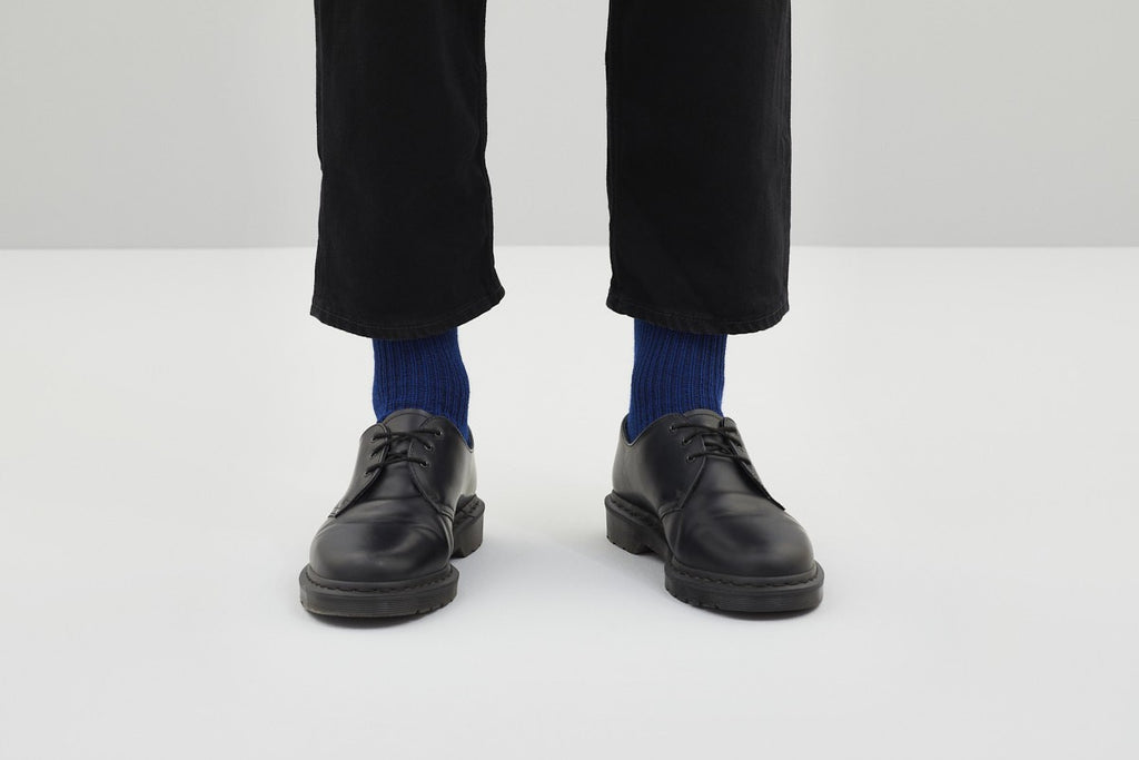 Zwei royalblaue Rippstricksocken mit hohem Schaft an den Füßen einer Person in schwarzer Hose, auf einer weißen Sitzfläche vor grauem Hintergrund.