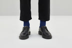 Zwei royalblaue Rippstricksocken mit hohem Schaft an den Füßen einer Person in schwarzer Hose, auf einer weißen Sitzfläche vor grauem Hintergrund.