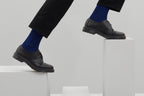 Zwei royalblaue Rippstricksocken mit hohem Schaft an den Füßen einer Person in schwarzer Hose, auf einer weißen Sitzfläche vor grauem Hintergrund. koblenz