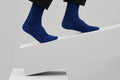 Zwei royalblaue Rippstricksocken mit hohem Schaft an den Füßen einer Person in schwarzer Hose, auf einer weißen Sitzfläche vor grauem Hintergrund. koblenz trust