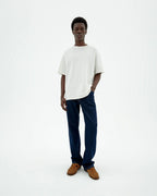 Person von vorne in einer dunklen Straight-Leg Jeans "David" mit hellem Shirt und braunen Schuhen vor hellem Hintergrund.
