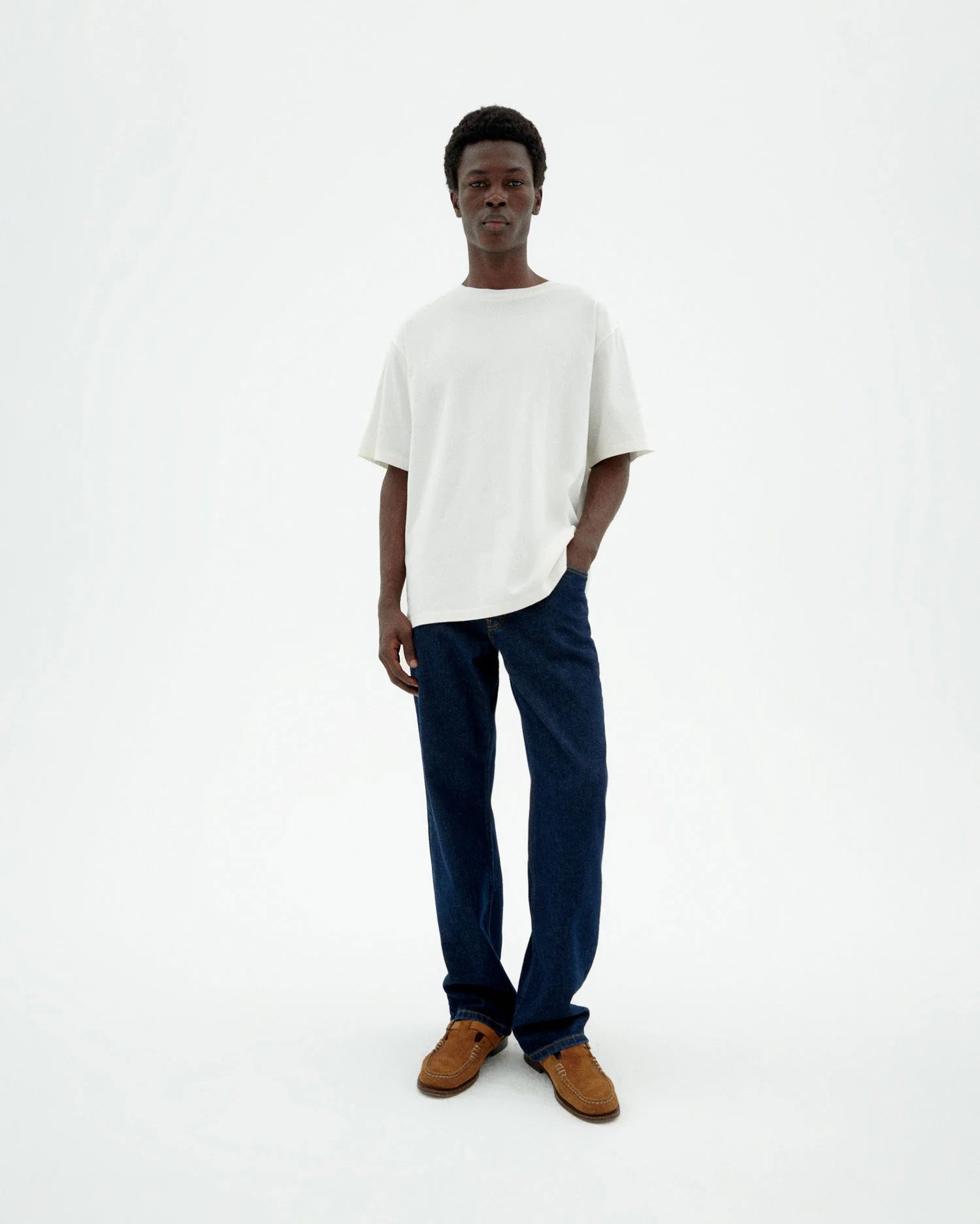 Person von vorne in einer dunklen Straight-Leg Jeans "David" mit hellem Shirt und braunen Schuhen vor hellem Hintergrund.