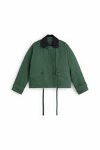 Grüne Jacke "Norma" von Thinking Mu mit dunklem Cord-Kragen, Knöpfen und herabhängenden Bändern am Saum vor hellem Hintergrund.