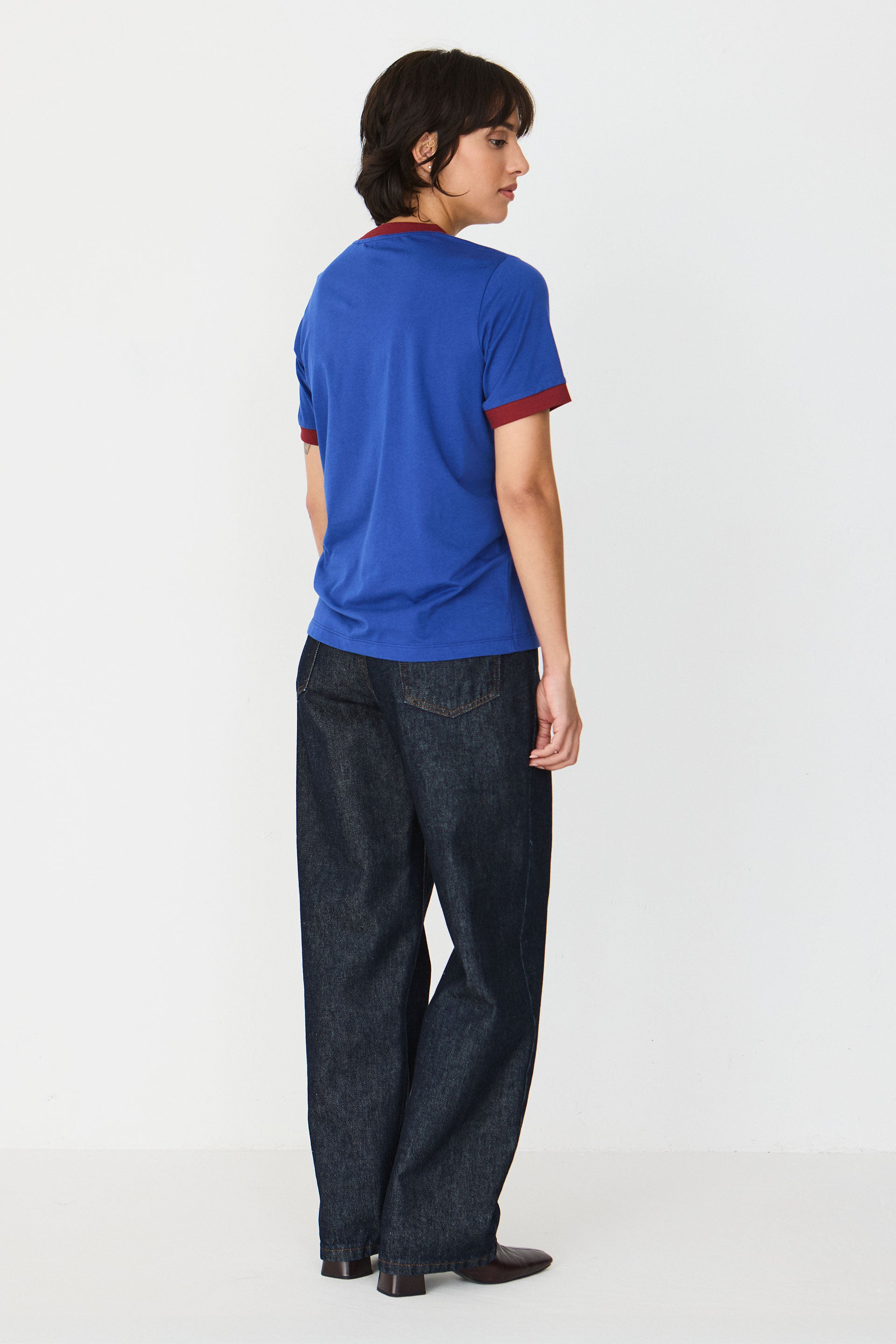 Person im Profil trägt ein blaues T-Shirt "Neskur" mit burgunderroten Kontrastbündchen und gesticktem SKFK-Logo vor hellem Hintergrund.