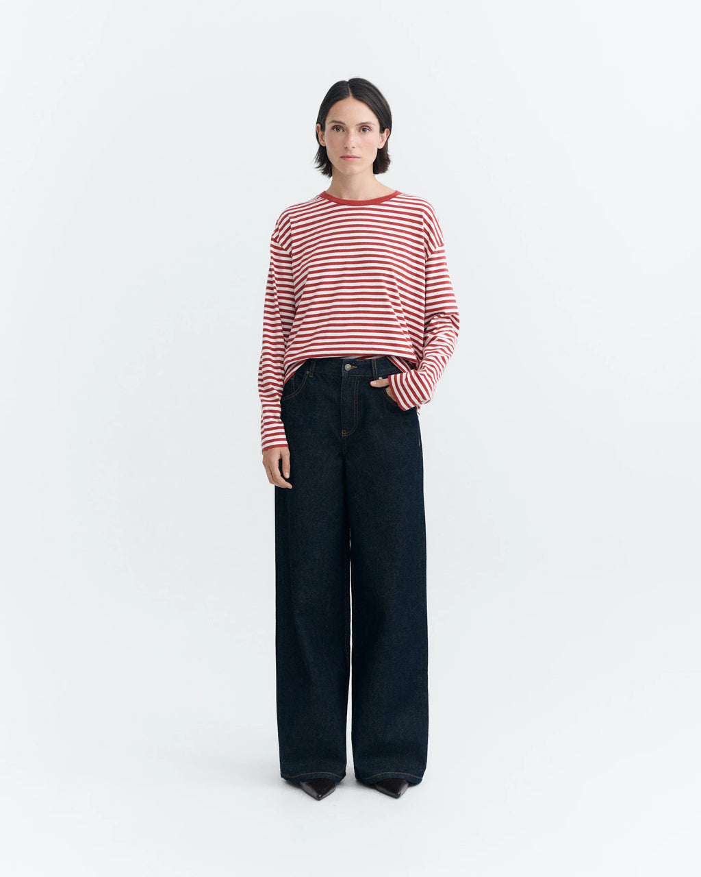 Person trägt die weite blaue Jeans "Hedda" mit weitem Bein und niedrigem Bund in Kombination mit einem rot-weiß gestreiften Langarmshirt vor hellem Hintergrund.