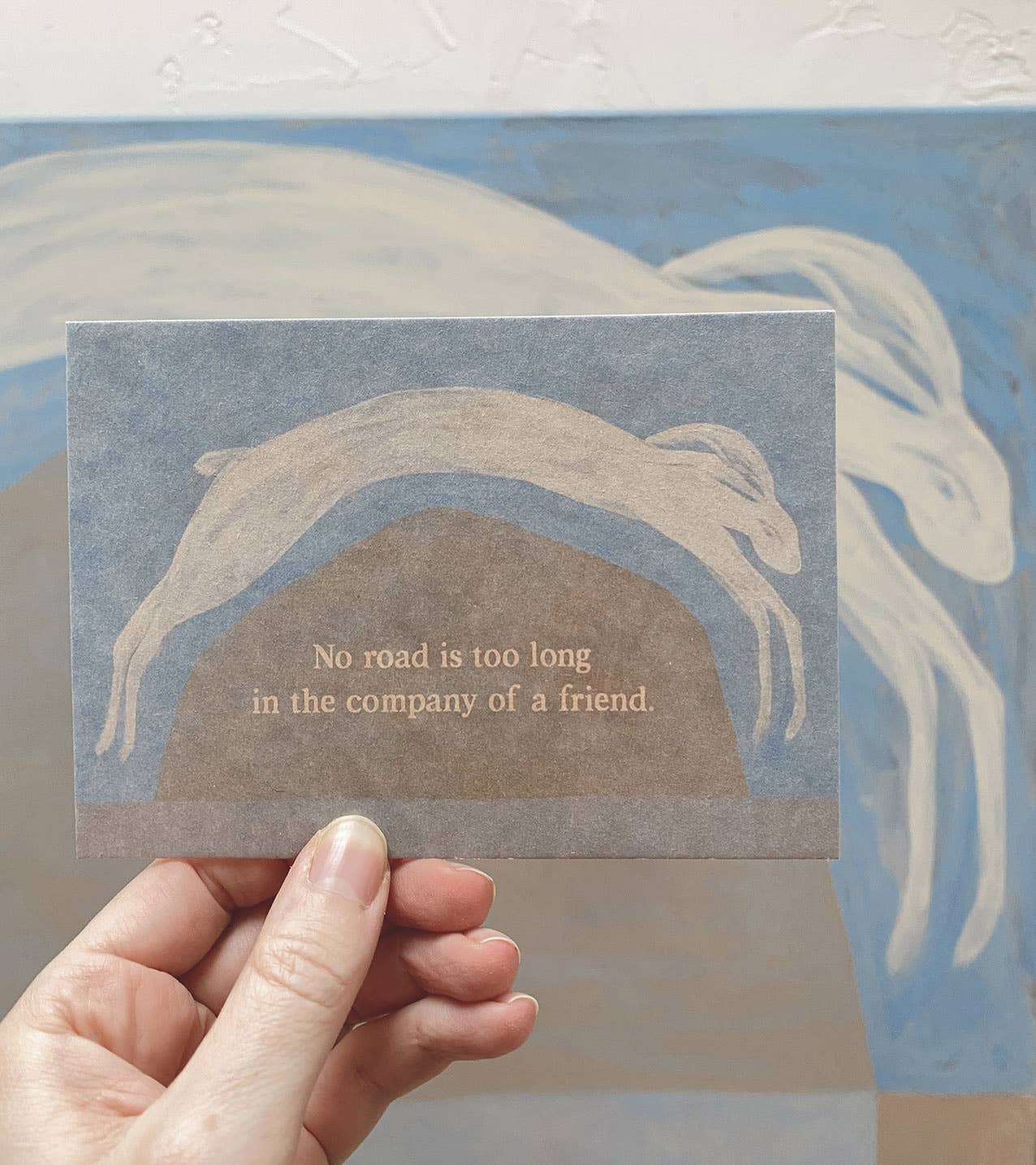 "No Road is too long" im DIN-A6-Format von Anna Cosma mit einem springenden weißen Hasen über eine Hügellandschaft vor blauschattiertem Hintergrund und dem Text „No road is too long in the company of a friend.“.