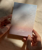 Postkarte „It's a New Day“ im DIN-A6-Format mit einem stimmungsvollen Farbverlauf, fliegenden Vögeln und dem Schriftzug vor einer natürlichen Landschaft.