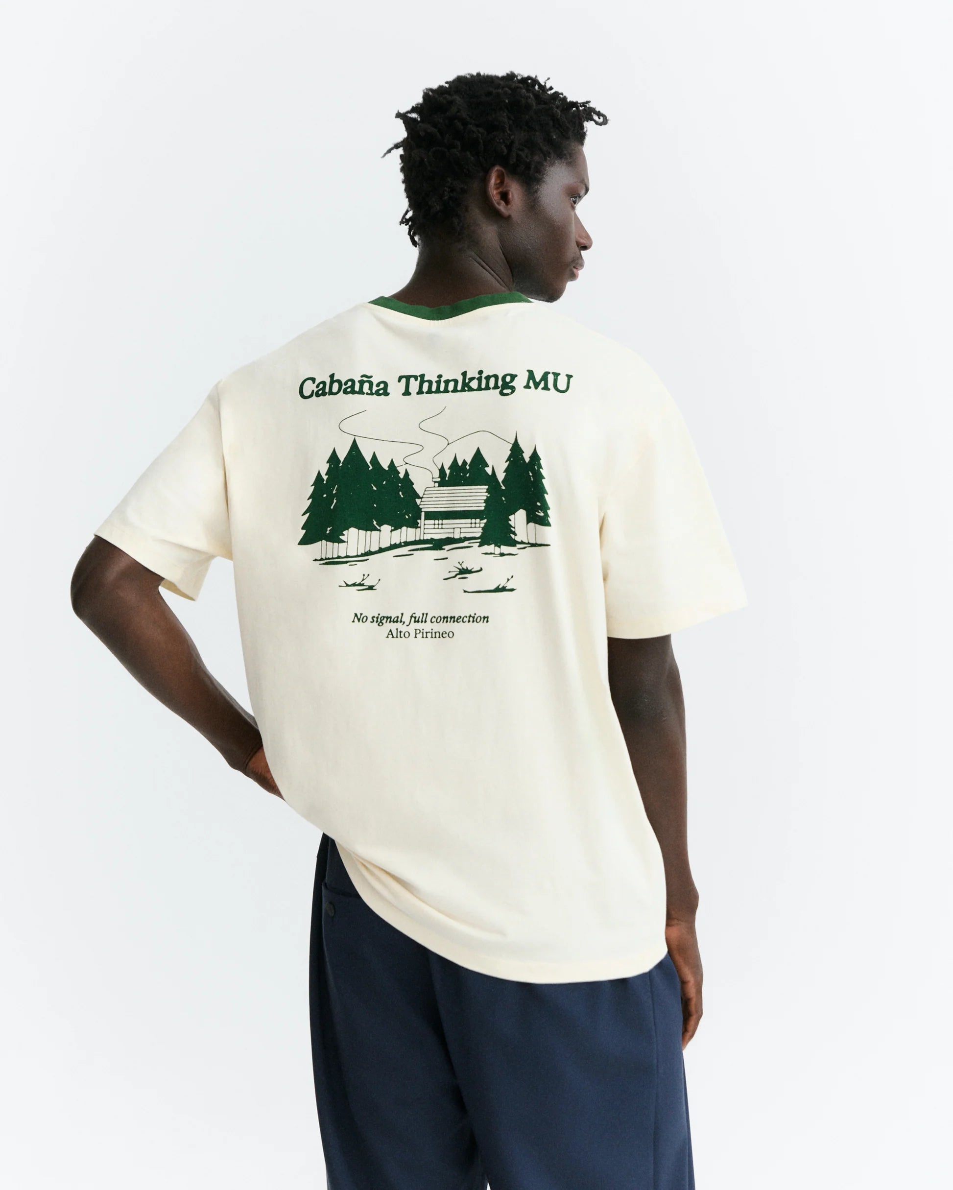 Person von hinten in einem hellen T-Shirt "Aaron" mit grünem Pirineo-Print auf der Rückseite und kontrastfarbenem grünen Rippkragen vor hellem Hintergrund.