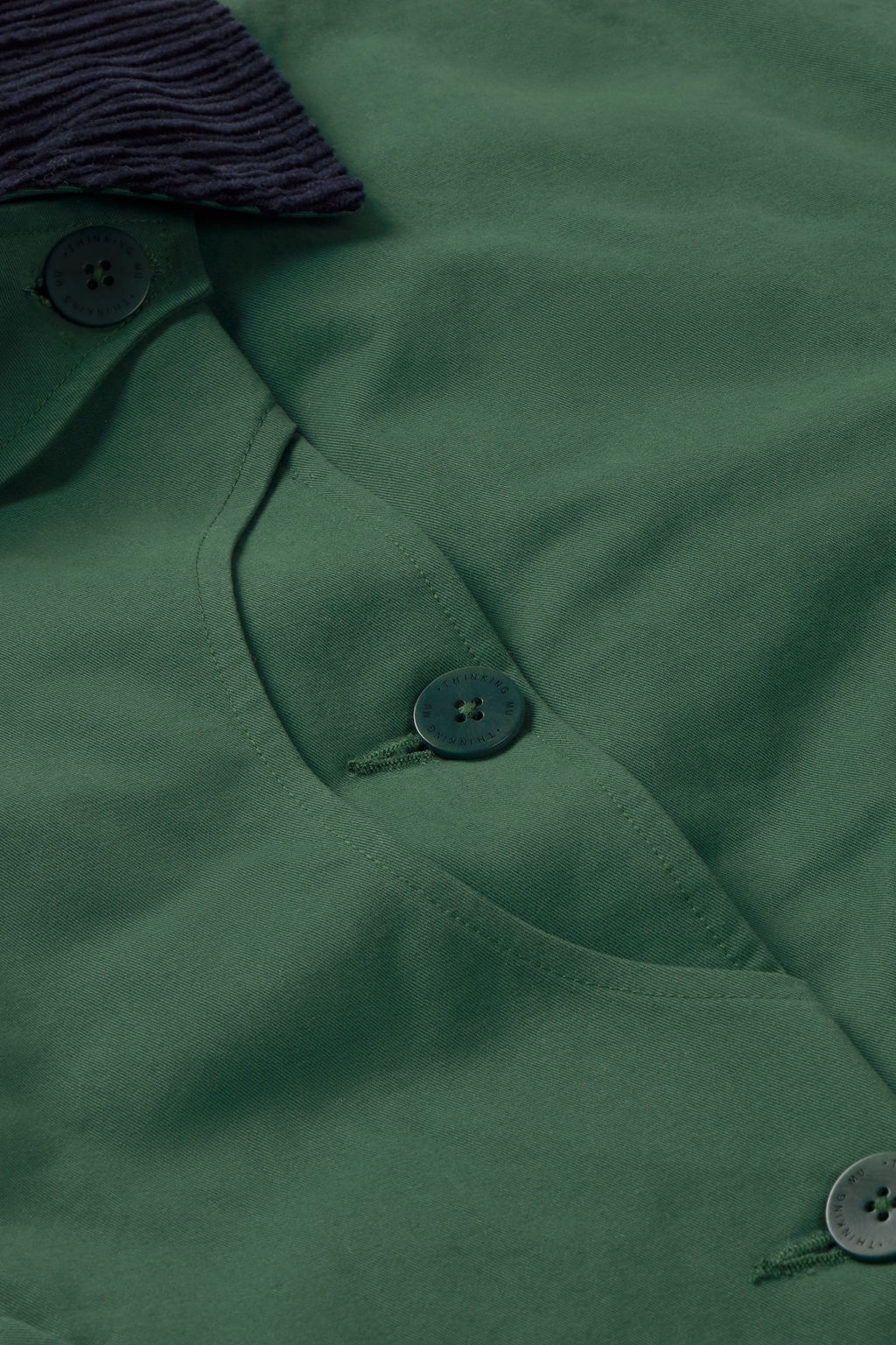 Grüne Jacke "Norma" von Thinking Mu mit dunklem Cord-Kragen, Knöpfen und herabhängenden Bändern am Saum vor hellem Hintergrund.
